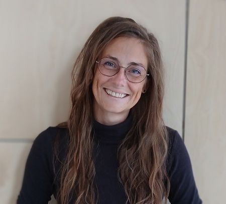 Zu sehen ist eine junge Frau mit langen braunen Haaren und Brille und schwarzem Pullover, die mit schräg gelegtem Kopf in die Kamera lächtelt
