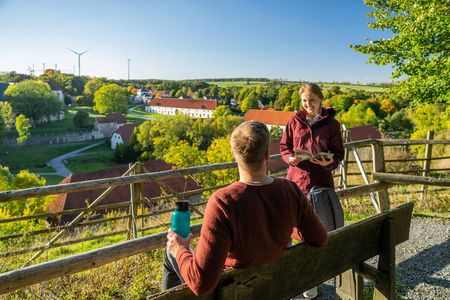Paar sitzt auf einer Bank mit Blick auf ein idyllisches Dorf in einer grünen, ländlichen Umgebung.