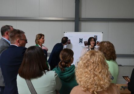 Eine Frau steht an einer Pinnwand und zeigt der umstehenden Gruppe ein Foto.