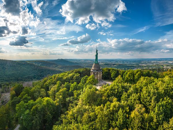 Eine Panorama-Ansicht des Hermanns-Denkmals. Unter der imposanten Hermannstatue erstrecken sich grüne, bewaldete Berge, über dem erhobenen Schwert erstreckt sich ein blauer Wolkenhimmel.