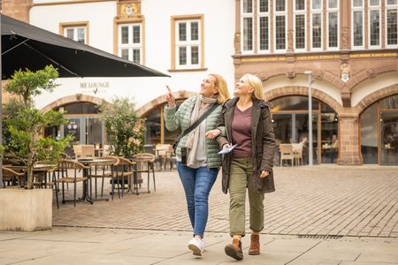 Zwei lächelnde Frauen spazieren Arm in Arm auf einem gepflasterten Platz, umgeben von historischen Gebäuden.