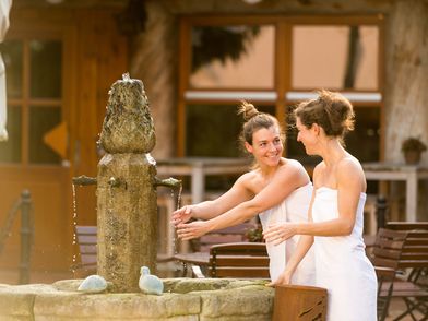 Zu sehen sind zwei Frauen in Handtüchern, die ihre Hände in einer Sauna-Outdoor-Landschaft im Brunnen waschen.