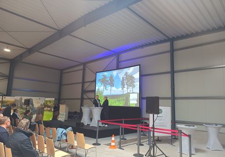 Das Bild zeigt eine Bühne und Leinwand; auf der Bühne ist der Moderator im Gespräch mit einer weiteren Person.