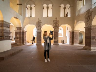 Zu sehen ist eine Frau mit Tablet in der Hand, die inmitten eines alten Kirchenraumes steht. Das Tablet hält sie hoch in Richtung Wand und betrachtet etwas dadurch. 