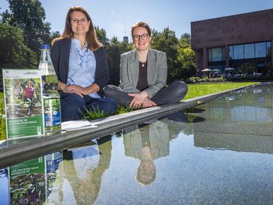 Zwei lächelnde Frauen sitzen im Schneidersitz, vor Ihnen Broschüren auf dem Rand eines Wasserbeckens, hinter ihnen die Kunsthalle Bielefeld