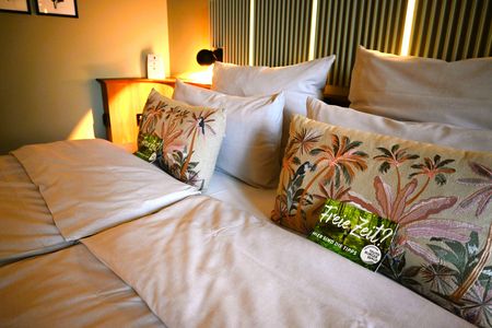 Zu sehen ist ein Hotelzimmer, auf den Betten, an die Kissen angelegt, liegen attraktiv gestaltete Postkarten, auf jeder Seite eine.
