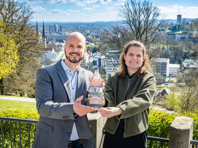 Zu sehen sind ein Mann und eine Frau, die gemeinsam lächelnd eine Mineralwasserflasche aus Glas mit einem auffälligen Familienfoto drauf in die Kamera halten. Im Hintergrund sieht man das Panorama der Stadt Bielefeld und die Sparrenburg.