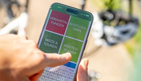 Hand hält Smartphone und Finger zeigt auf Display, darauf zusehen ist der Teuto-Navigator