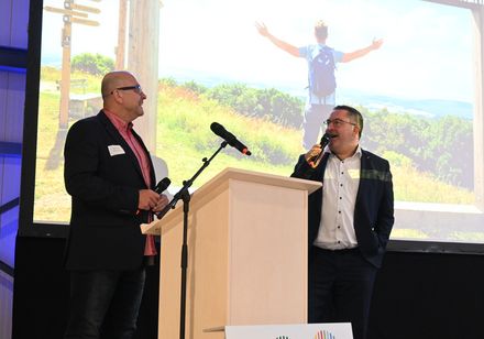 Man sieht eine Leinwand und eine Bühne auf der zwei Menschen im Gespräch sind.