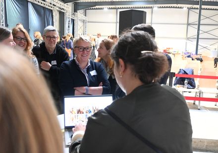 Eine Frau steht vor einem aufgeklappten Laptop, eine Gruppe steht vor ihr.