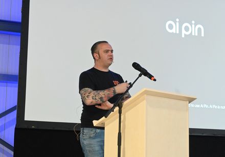 Ein Mann steht mit einem Mikro auf einer Bühne vor einer Leinwand und hält einen Vortrag.