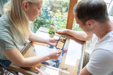 Eine Frau zeigt einem Mann ein Smartphone in einem stilvollen Café mit Holzdekor und Blumen.
