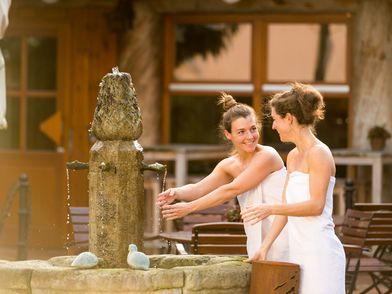 Zwei Frauen stehen nur mit Handtuch bekleidet in einem Saunagarten und lächeln sich an. Eine wäscht sich die Hände in einem Brunnen.