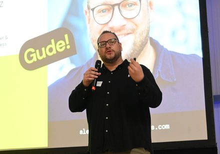 Ein Mann steht mit einem Mikro vor einer bunten und hell erleuchteten Leinwand und hält einen Vortrag.