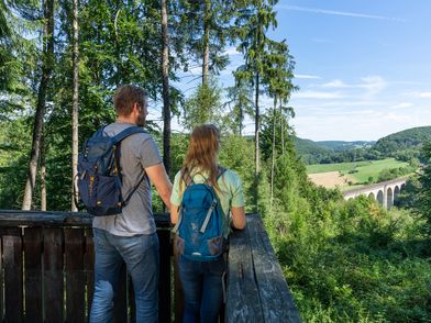 Zu sehen sind ein Mann und eine Frau von Hinten, mit Rucksäcken, die auf das Viadukt im Paderborner Land blicken
