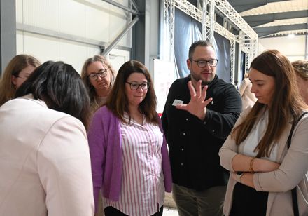 Eine Gruppe steht mit einem Referenten zusammen; der Referent erläutert einen Sachverhalt.