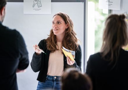 Eine Frau steht mit Materialien in der Hand in einer Workshopatmosphäre vor den Teilnehmenden.