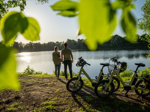 Der Betrachtende sieht durch die Blätter eines Baumes das Wasser eines großen Sees im Sonnenlicht funkeln. Am Rufer, im rechten unteren Bildrand, stehen zwei Fahrräder, direkt am Rand des Sees steht ein Paar und blickt aufs Wasser.