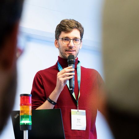 Zu sehen ist ein junger Mann mit braunen kurzen Haaren und Brille und einem roten Pullover, der in ein Handmikrofon spricht