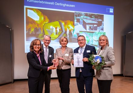 Auf einer Veranstaltung überreichen eine Frau und ein Mann zwei weiteren Frauen und einem Mann einen Preis.