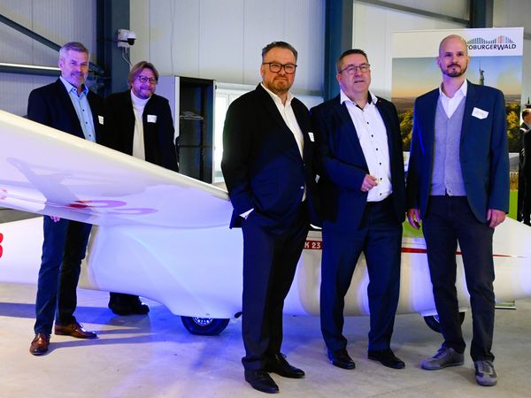 Zu sehen sind fünf männliche Personen in Anzügen, die sich rund um ein kleines Segelflugzeug aufgestellt haben.