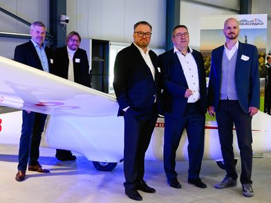 Zu sehen sind fünf männliche Personen in Anzügen, die sich rund um ein kleines Segelflugzeug aufgestellt haben.