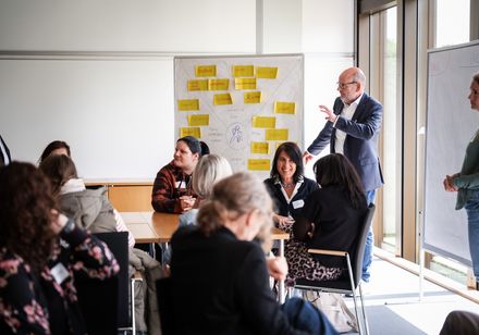 Ein Workshopleiter steht vor einer Pinnwand und erläutert den Teilnehmenden etwas.