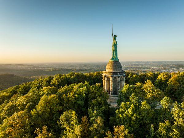Zu sehen ist das Hermannsdenkmal, das auf einem Berg aus den grünen Baumkronen ragt. Im Hintergrund sieht man Landschaft und einen blauen Abendhimmel