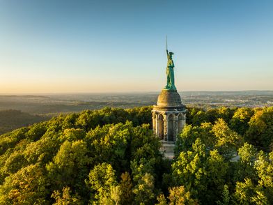 Zu sehen ist das Hermannsdenkmal, das auf einem Berg aus den grünen Baumkronen ragt. Im Hintergrund sieht man Landschaft und einen blauen Abendhimmel