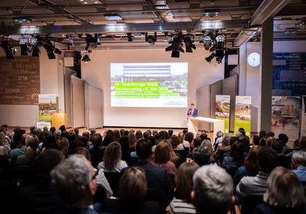 Ein Moderator steht vor einer Leinwand und begrüßt die Teilnehmenden zur Veranstaltung. 