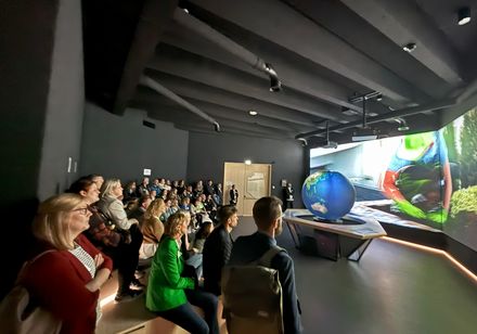 Rechts im Bild befindet sich eine Leinwand, davor die Nachbildung der Erde; im davor gelagerrten dunklen Zuschauerraum sitzt eine Gruppe.