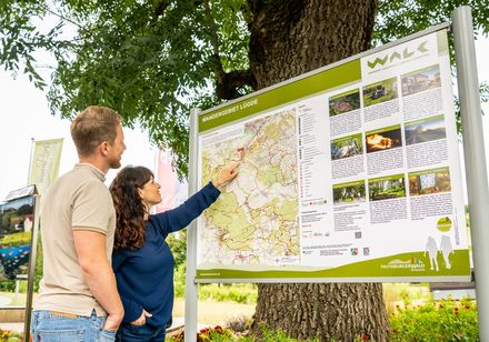 Ein Paar betrachtet eine Wanderkarte an einem Infopunkt im Freien, umgeben von grüner Natur.
