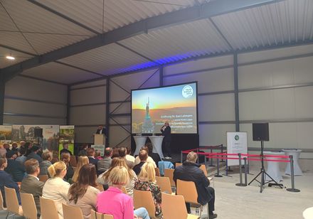 Zuschauer sitzen in Reihen vor einer Bühne und einer Leinwand, die das Hermannsdenkmal zeigt.