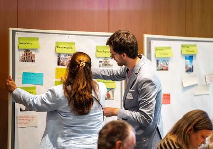 Eine Frau und ein Mann stehen in einer Workshopatmosphäre vor einer Pinnwand.