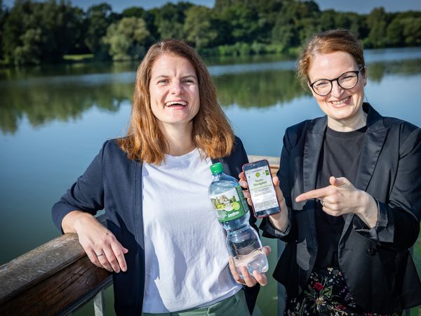 Zwei Frauen stehen vor einem See und halten lächelnd eine Carolinen Mineralwasserflasche mit Sonderetikett und ein Handy mit Erlebnis-App in die Kamera.