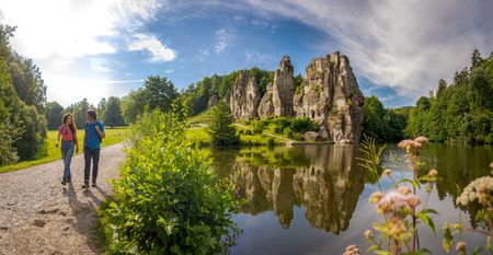 Zwei Personen wandern entlang eines Sees, dessen Wasser malerisch die markanten Externsteine spiegelt.