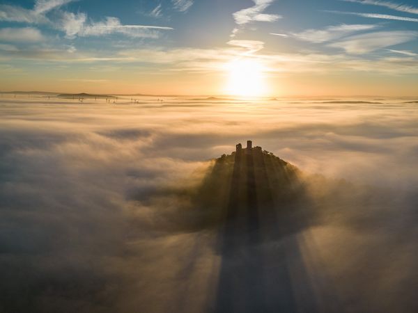 Ein Hügel mit einer Burgruine ragt aus dem Nebenmeer. Im Hintergrund färbt die Abendsonne den Himmel. 