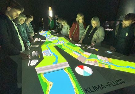 Eine Gruppe von Menschen betrachtet in einem Museum eine hell erleuchtete Informationstafel.