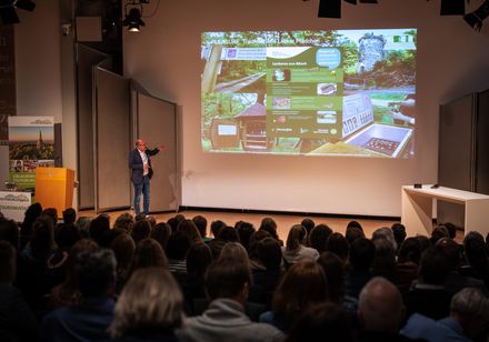 Ein Mann hält einen Vortrag vor Publikum und deutet auf seine Präsentation an der Leinwand.
