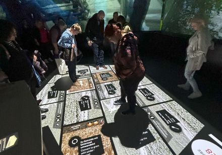 In einem verdunkelten Raum betrachten Museumsbesucher interaktive Informationstafeln am Boden.